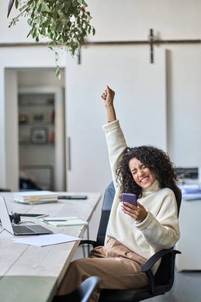 Euphoric young female worker holding mobile phone celebrating win. Euphoric young female worker holding mobile phone celebrating win receiving good news about job promotion, getting hired, feeling happy, rejoicing success with yes reaction working in office. Vertical incentive stock pictures, royalty-free photos & images