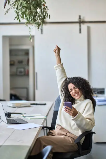 Euphoric young female worker holding mobile phone celebrating win receiving good news about job promotion, getting hired, feeling happy, rejoicing success with yes reaction working in office. Vertical