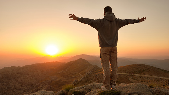 Man standing with open arms on a mountaintop. Person standing on the cliff edge facing the rising sun. Scenery sunrise in mountainside. Embracing the world