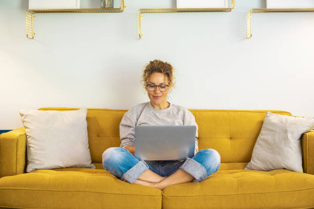 istock One woman smiling and using laptop computer at home sitting comfortably on a yellow sofa in living room. Smart working female people notebook. Surfing the web. Enjoying technology and connection 1463688568