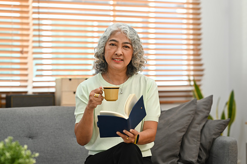 Smiling middle aged woman reading book on couch, enjoy stress free peaceful mood wellbeing in comfortable home.