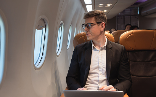 Businessman in formal wear and glasses looking out of aircraft window during flight  while working with laptop computer. Internet connection on board service and business travel concept