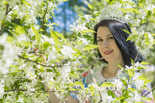 A young beautiful brunette girl in a summer blue dress and with a diadem on her head stands among the flowers of the apple tree. Good spring mood.
