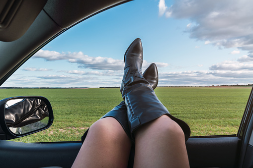 Crossed legs of a Caucasian woman in cowboy boots out of the car window, sunny day with clouds