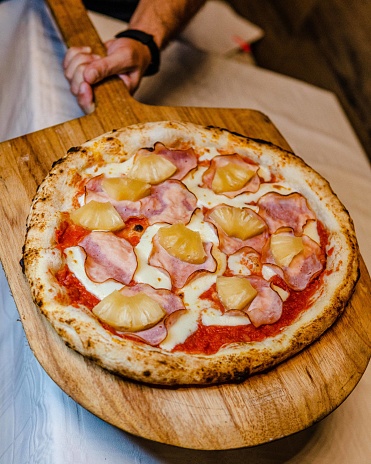 A man's hand holding a wooden board with a crunchy pizza with ham and pineapple