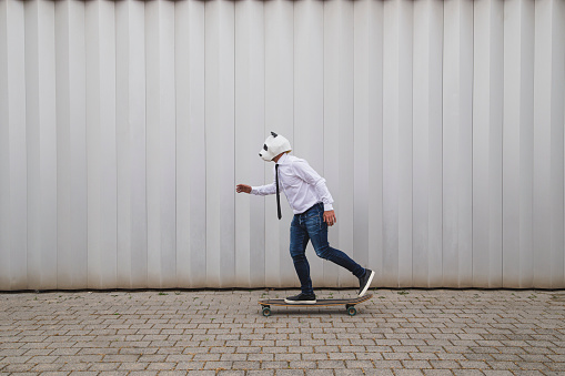 A male in a panda head mask skateboarding on a longboard against a gray wall