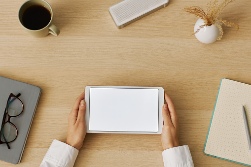 Minimal top view of female hands holding digital tablet with white screen mockup at desk workplace, copy space