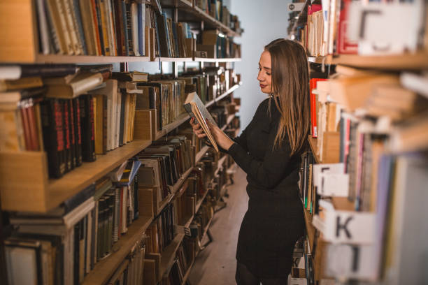 istock Side view of a smiling librarian reading a book while standing leaning against bookshelf in library 1463778404