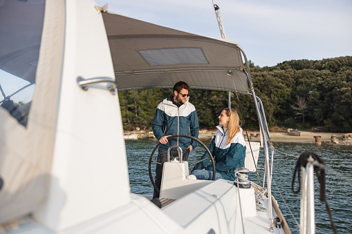 Three-quarter shot with blurred background and left side of a young Caucasian couple looking at each other and matching jackets while sailing on a summer day at the Adriatic sea.