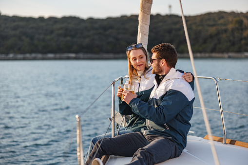 Three-quarter side shot with blurred background of a happy young Caucasian couple toasting with beer while seated on the bow of a sailing boat and enjoying the stunning Adriatic sea view on a summer day.