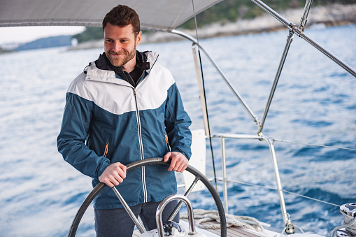 Three-quarter side shot with defocused background of a good looking sailor slightly smiling and looking at the horizon while holding firmly the boat helm enjoying his job on summer day in the Adriatic sea.