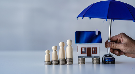 Blue umbrellas on houses and small cars, stacks of money, wooden dummy, protection, insurance industry, real estate Property security, protection concept under blue umbrella isolated on white