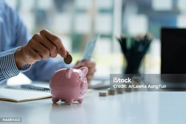 Hands Of A Young Asian Businessman Man Putting Coins Into Piggy Bank And Holding Money Side By Side To Save Expenses A Savings Plan That Provides Enough Of His Income For Payments Stock Photo - Download Image Now
