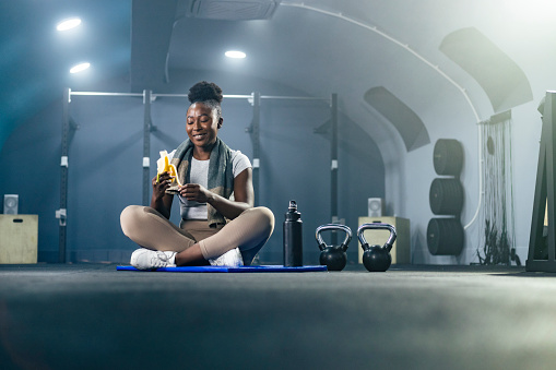 Fit African American woman in sportswear eating banana at the gym after workout. She is sitting on yoga mat.
