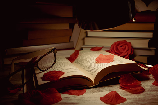 open book with glasses on top with rose petals scattered on top and books piled up in the background.
