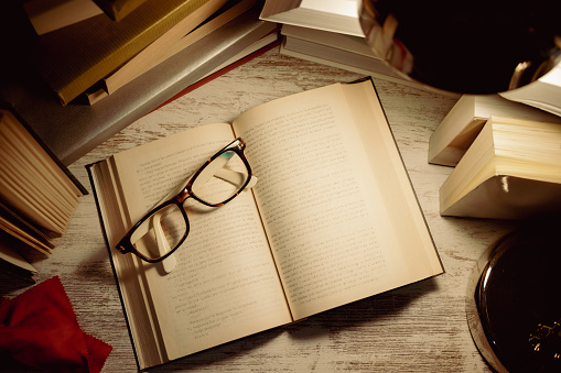 top view open book with glasses on top surrounded by stacked books and the light from a lamp