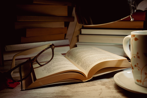 open book with a pair of glasses on top of it in the light of a lamp, a cup and in the background books piled up