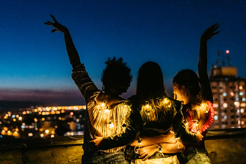 Rear-view of three beautiful young female friends coverwith string lights at night on the rooftop.