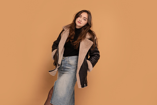 Trendy young brunette woman with long hair wearing an autumn coat and jeans skirt, posing over studio background. A lot of copy space. Fashionable girl.