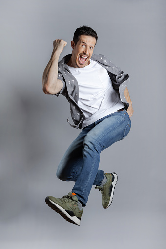 Young overjoyed caucasian man wearing casual white shirt jumps high with winner gesture, Full-length, studio shot, isolated