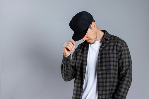 Young man in casual clothes, white t-shirt checkered shirt looking down while touching his cap, hidden face, posing isolated on gray background, studio portrait. Horizontal