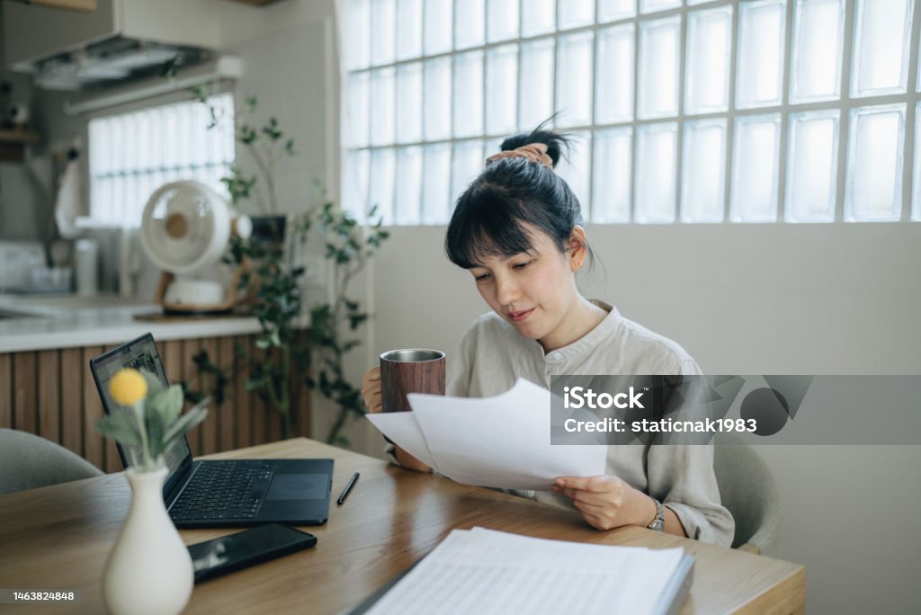 Asian women working with invoices, calculates expenses doing work at home. Young focused woman in spectacles calculating budget at home, sitting at table with laptop, holding paper taxes bills and reading financial information. Individuality Stock Photo