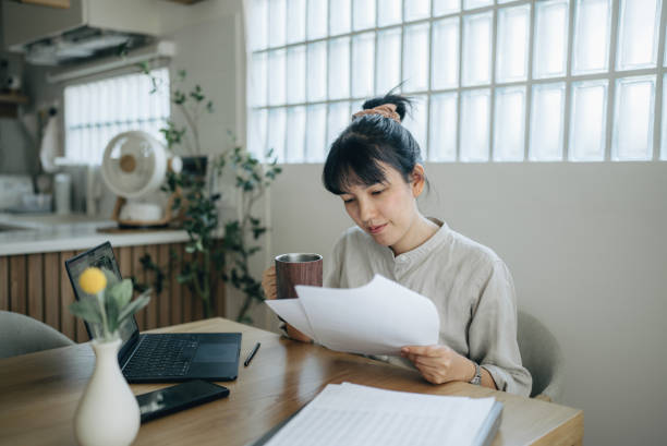 Asian women working with invoices, calculates expenses doing work at home. Young focused woman in spectacles calculating budget at home, sitting at table with laptop, holding paper taxes bills and reading financial information. personal loan stock pictures, royalty-free photos & images