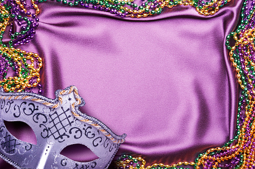 Mardi Gras mask and beads and a carnival mask on a purple satin background.
