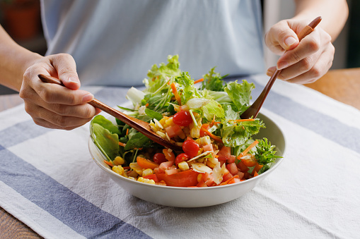 Cropped image asian woman mixing ingredients in her healthy fresh vegan salad. A multi-coloured healthy salad bowl with assorted fresh fruits and vegetables, lettuce, tomatoes, carrot and corn close-up