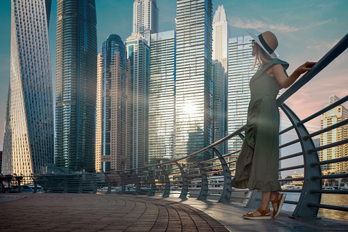tourist woman in fashionable summer dress and hat enjoying Dubai marina skyline at sunset. copy space