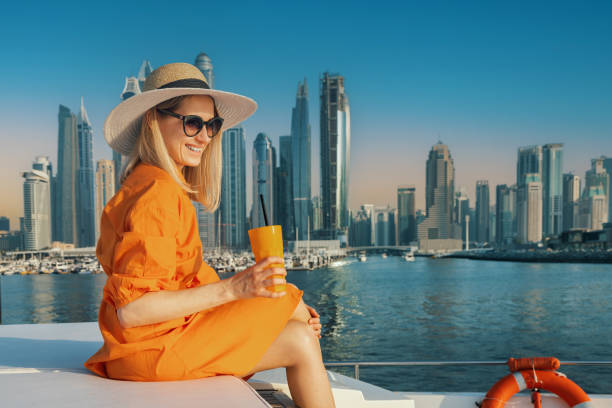 donna sorridente in vestito arancione con drink su yacht di lusso a dubai al tramonto. skyline della città sullo sfondo. copia spazio - dubai yacht luxury marina foto e immagini stock