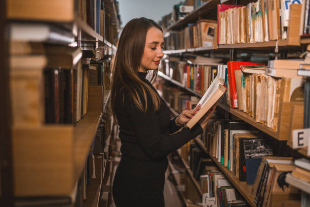 istock Side view of lovely librarian reading a book while standing leaning against bookshelf in library 1463847210