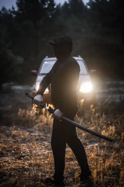 In the headlights of a car in the forest, a silhouette of a man with a sword in his hands is visible. At dusk in the rain, a man trains with a sword in the forest..
