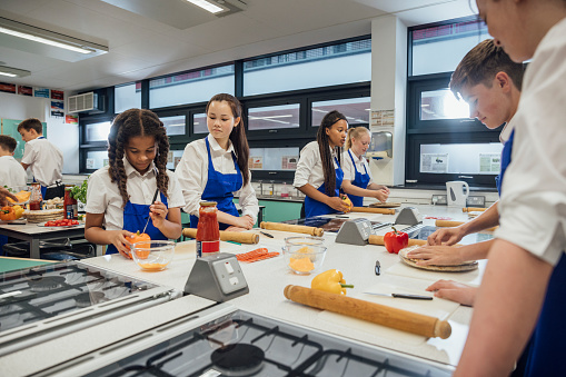 A low-angle view of a group of high school students working in pairs and small groups to make their own fresh pizza from scratch. They have been given safety instructions on how to cook safely.