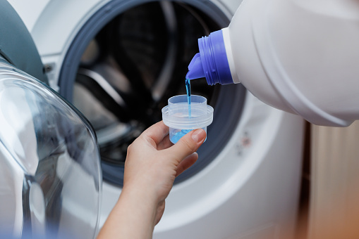 Close up shot of a woman hands squeezing liquid gel laundry detergent from a plastic bottle into a small container, with a washing machine in the background