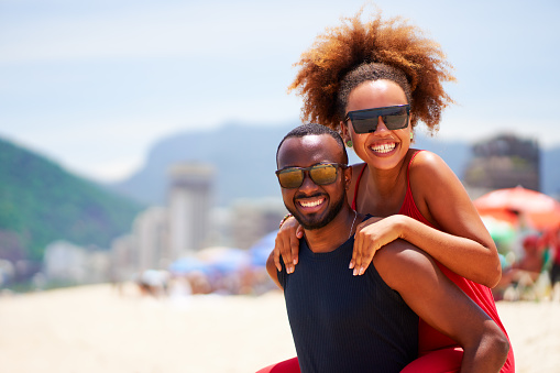 portrait young black brazilian carioca couple in piggyback position smiling and having fun in Ipanema beach Brazil