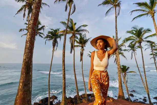 Photo of Young african female model posing in colorful clothes at tropical location at sunrise. Black woman against exotic scenery at dawn. Multiracial dark-skinned model poses in front of palm trees at sunset