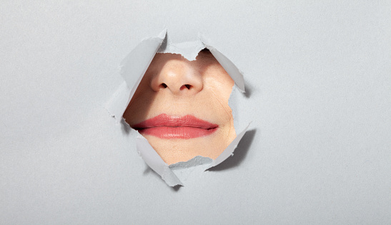 Woman showing her lips playfully in torn paper hole, through breakthrough of gray background. Cosmetics advertising concept. Closeup