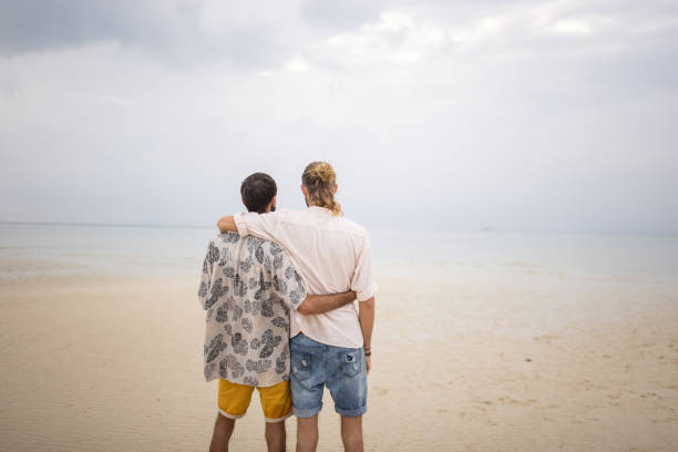 Two men, happy gay couple standing on the beach by the sea in Thailand.