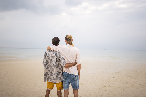 Two men, happy gay couple standing on the beach by the sea in Thailand.