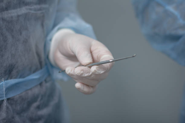 chirurg trzyma łyżkę volkmanna w wyciągniętej dłoni w rękawiczce medycznej na tle niebieskiego munduru. pielęgniarka trzyma w ręku narzędzie chirurgiczne podczas operacji. - surgeon isolated paramedic operating room zdjęcia i obrazy z banku zdjęć