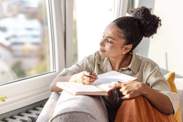 Young African American woman writing notes A young woman is at home, she is sitting on the sofa in the living room and writing notes diary stock pictures, royalty-free photos & images