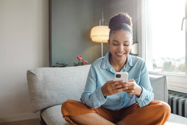 A young beautiful woman using a smart phone at home A young woman is at home in the living room, she is using a mobile phone and smiling home shopping stock pictures, royalty-free photos & images