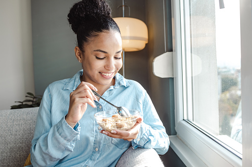 A young woman is eating breakfast at home by the window and enjoying