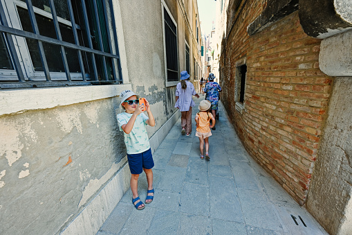 Kids walk narrow brick streets in Venice, Italy.  Boy tourist making photo by phone.