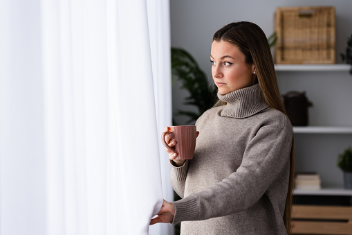 Winter clothing pretty woman looking through the window at home while holding cup of hot drink like tea or coffee