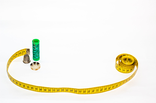Green thread coil with a pin nailed next to a yellow meter, a brown button and a metal thimble on a white background.