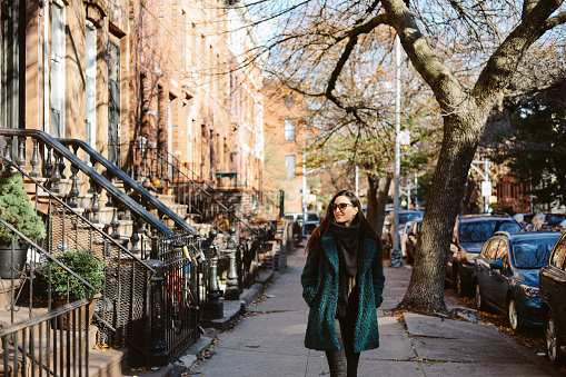 Young woman wearing a stylish fake fur coat walking in a beautiful Brooklyn neighbourhood in NYC, in-between the brownstone buildings.