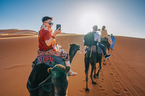 Asian Chinese Tourist Camel caravan going through the Sahara desert in Morocco at sunset