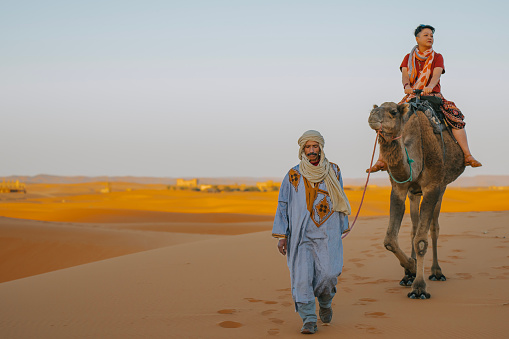 Asian Chinese Female Tourist riding Camel going through the Sahara desert in Morocco at sunset lead by camel driver